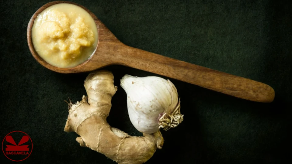 Picha inamuonesha kijiko cha mbao chenye mchanganyiko wa kitunguu saumu na tangawizi zilizosagwa na pembeni yake kuna kipande cha tangawizi na kitunguu saumu kimoja.